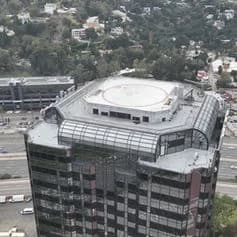 An aerial view of a tall building with a circular rooftop structure and glass facade, adjacent to a highway