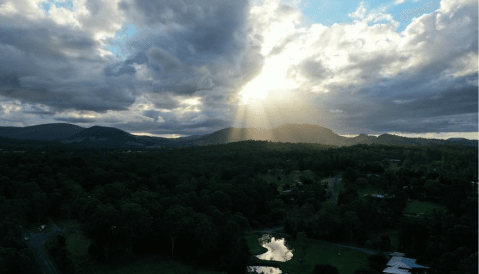 Aerial view of a forested landscape with hills, a small pond, and sun rays breaking through clouds