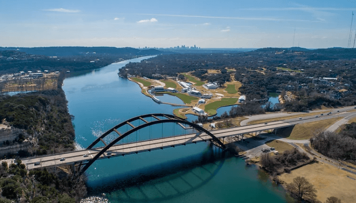 Aerial view of a curved arch bridge over a wide river, with a highway and green park along the banks