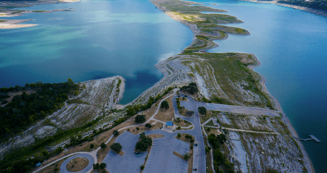 Aerial view of a winding peninsula along a blue water coastline with rocky slopes and a paved parking area