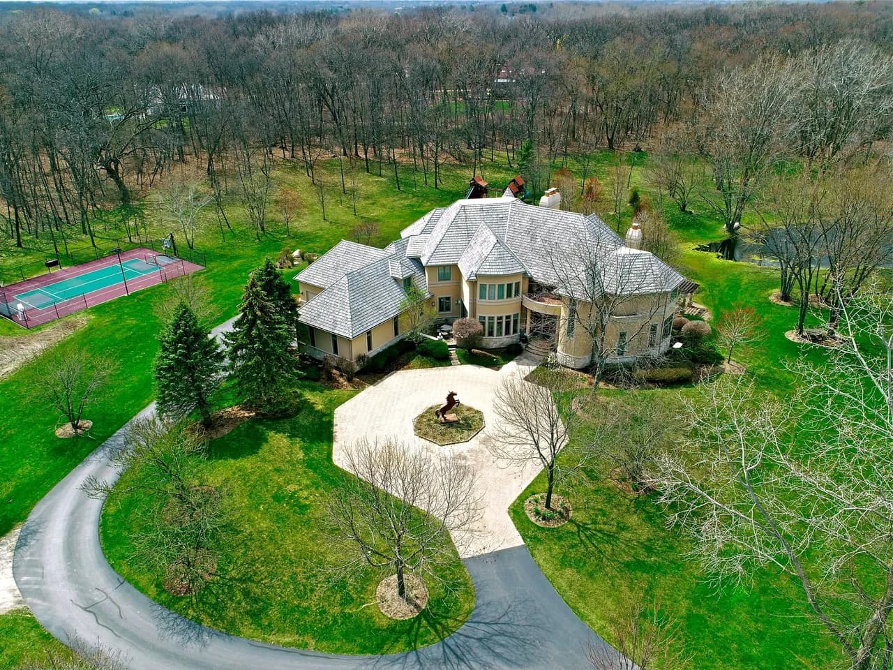 Aerial view of a large beige house with circular driveway, statue centerpiece, tennis court, and lawn