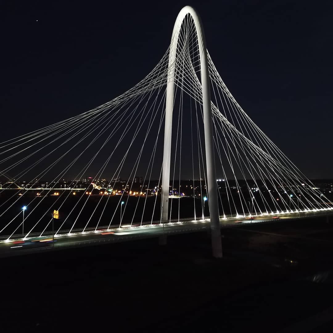 Night shot of a cable-stayed bridge with a tall white arch and lit cables, city lights in background