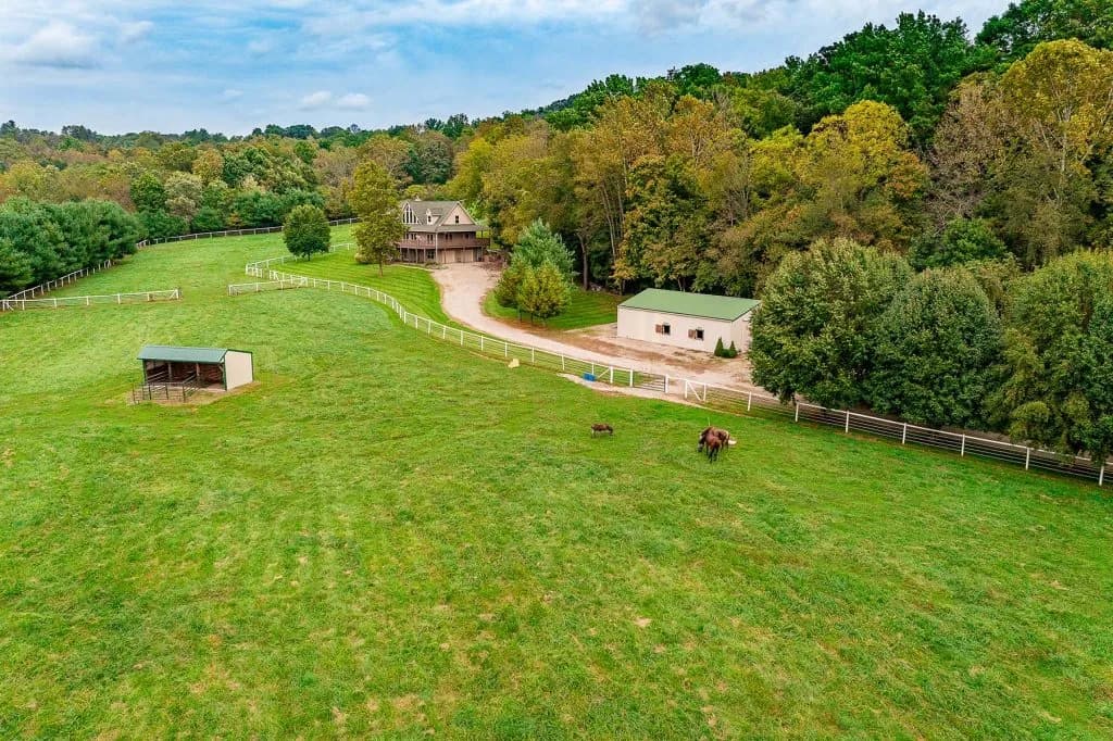 Aerial view of a rural farm with two horses in a fenced green field, buildings and trees