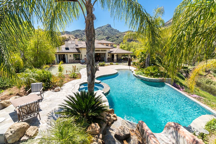 Turquoise pool with curved edges, stone patio, palm trees, outdoor seating, and a house in the background