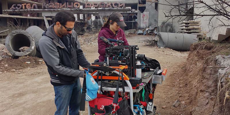 Two people stand beside a cart loaded with drone gear and bags in an industrial yard