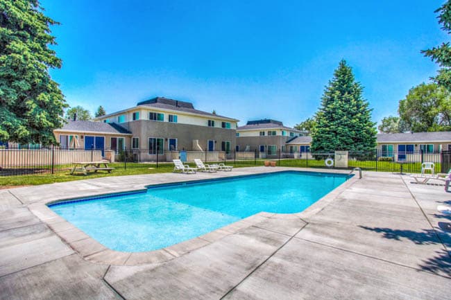 Blue rectangular pool with concrete deck, white lounge chairs, fenced yard and apartment buildings beyond