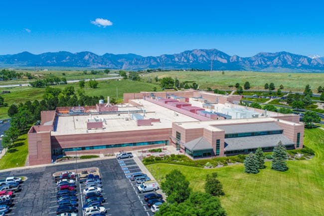 Aerial view of a large beige building complex with a parking lot, green fields, and distant mountains