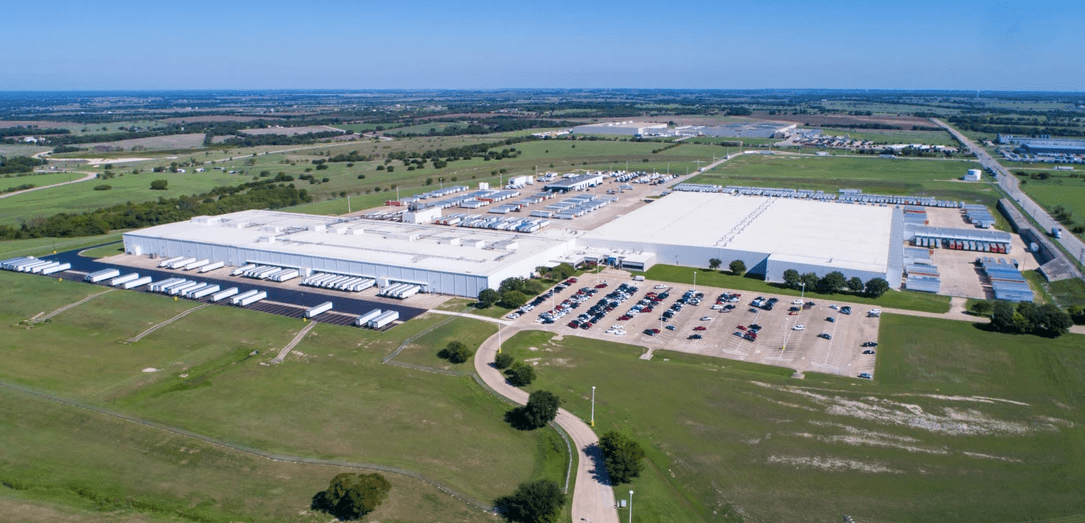 Aerial view of a large warehouse complex with loading docks, trailers, and a parking lot surrounded by fields