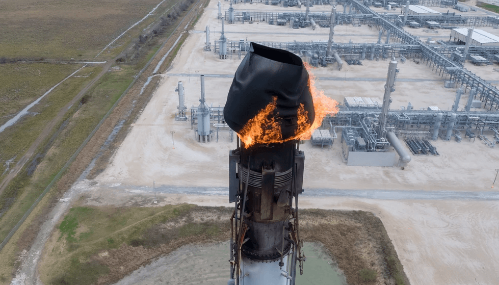 Flare stack with active fire atop a tall industrial tower, surrounded by processing facilities and roads