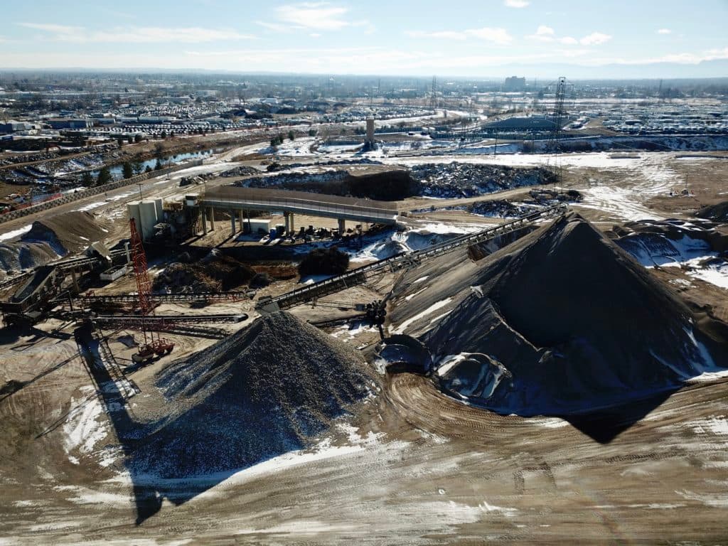 Aerial view of a mining site with large gravel piles, conveyors, a crane and snow on the ground