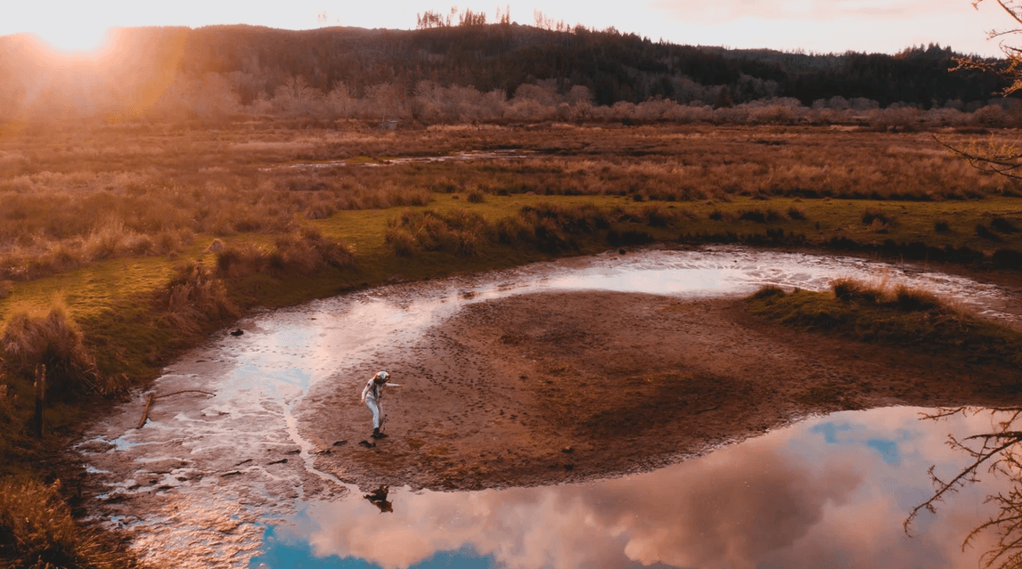 Aerial view of a person standing on the muddy shore around a circular pond in a marsh, sunset light