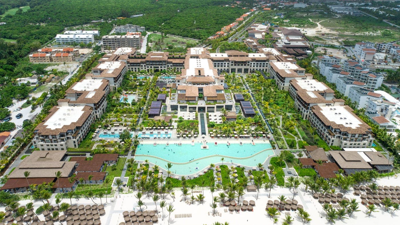 Aerial view of a resort with a central pool, palm trees, and U-shaped brown-roofed buildings near the beach