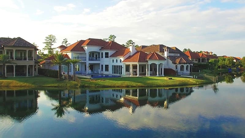 Row of white houses with red-tiled roofs along a lake, with palm trees and reflections in water