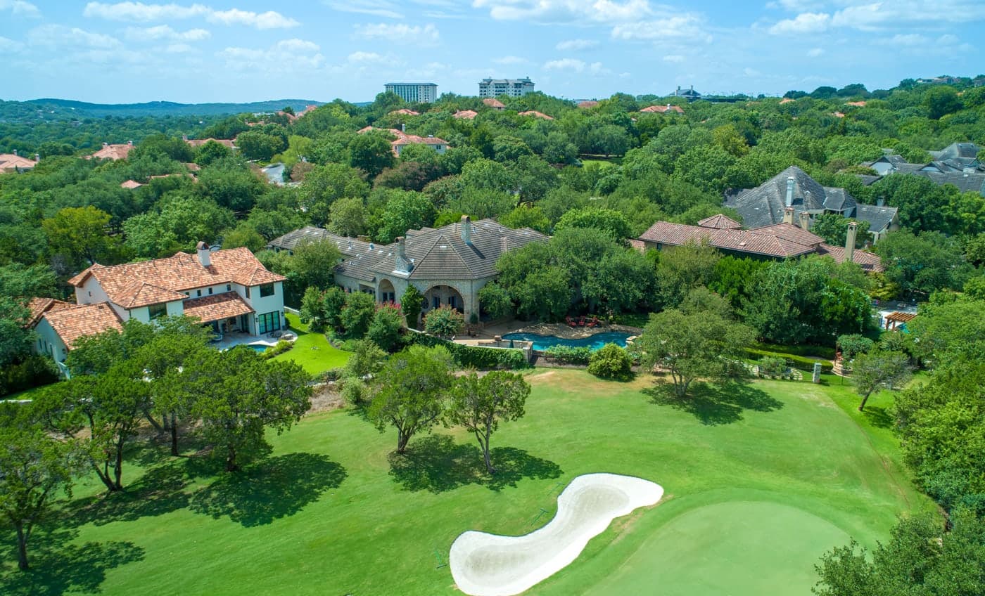 Aerial view of luxury homes with tile roofs, green lawns, trees, a pool, and a sand bunker
