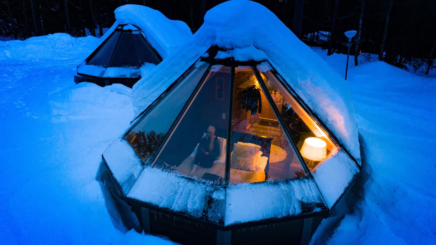 Two glass pyramid huts covered in snow, with blue lighting and warm interior light visible through the glass