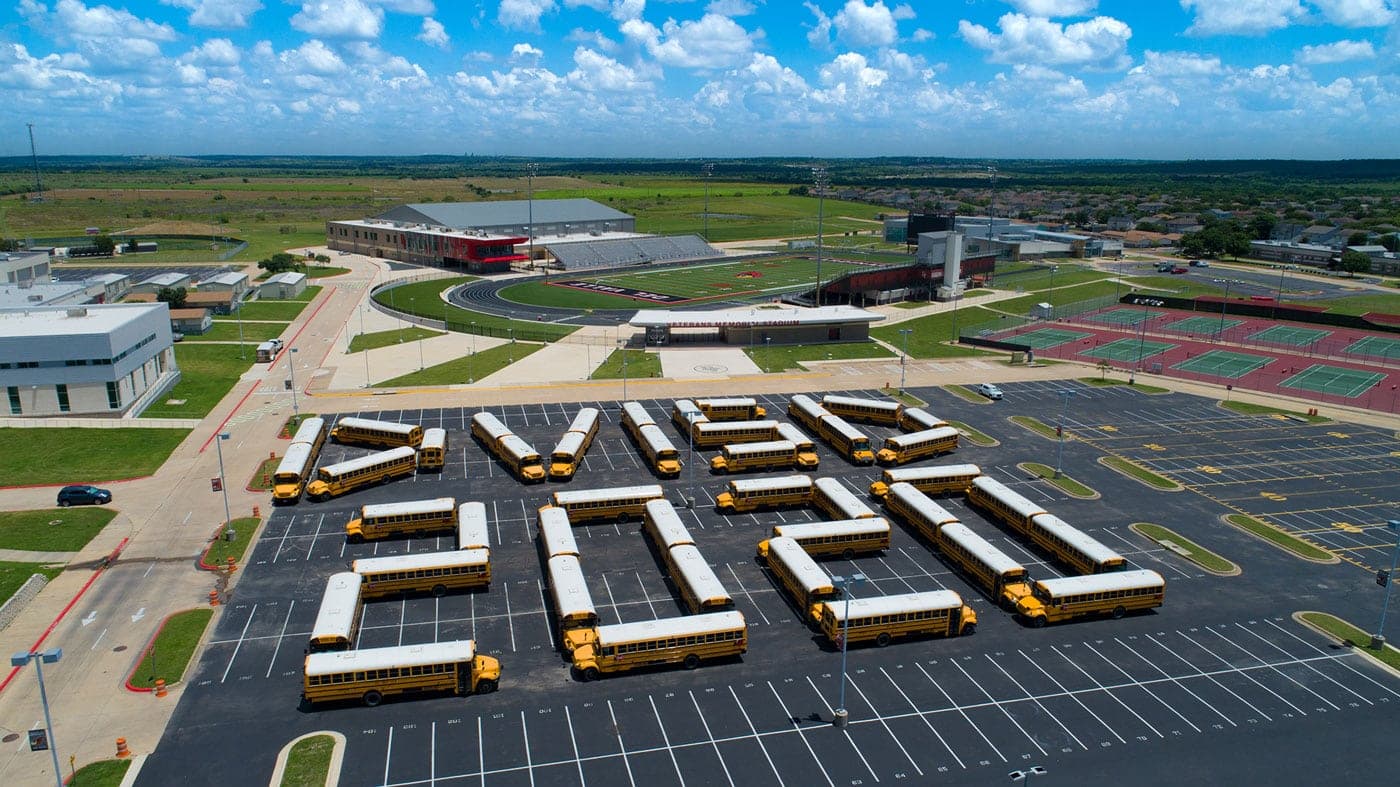 Aerial view of a school campus with a large parking lot where yellow buses are arranged to spell a word