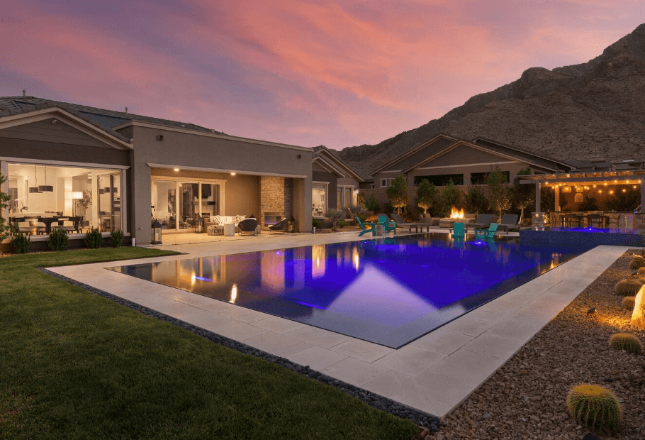 Backyard of a modern home with a rectangular blue-lit pool, outdoor seating, and desert mountains at sunset