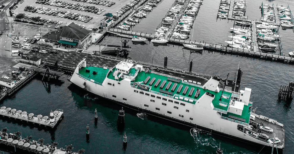 Aerial view of a large white ferry with a green top deck at a marina, with many boats and docks nearby