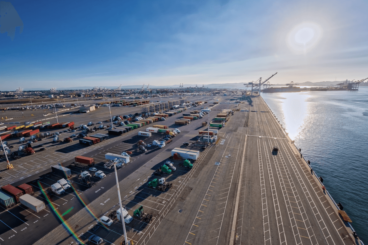Aerial view of a port with rows of shipping containers and trucks along a long pier by the water, sun glare