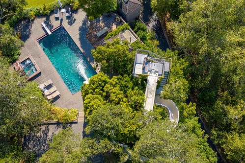 Aerial view of a backyard pool with turquoise water, a curved slide, surrounded by trees and a patio