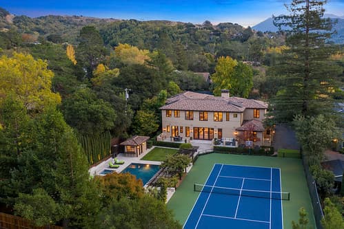 Aerial view of a two-story beige house with a brown roof, pool, and blue tennis court amid green trees