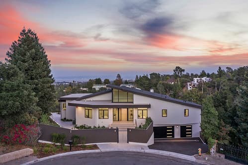 Aerial view of a modern white house with large windows and two-car garage, curved driveway, trees at sunset