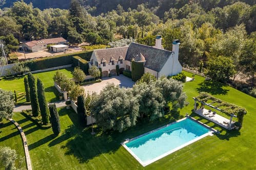 Aerial view of a white house with a gray roof, a manicured lawn and a rectangular turquoise pool