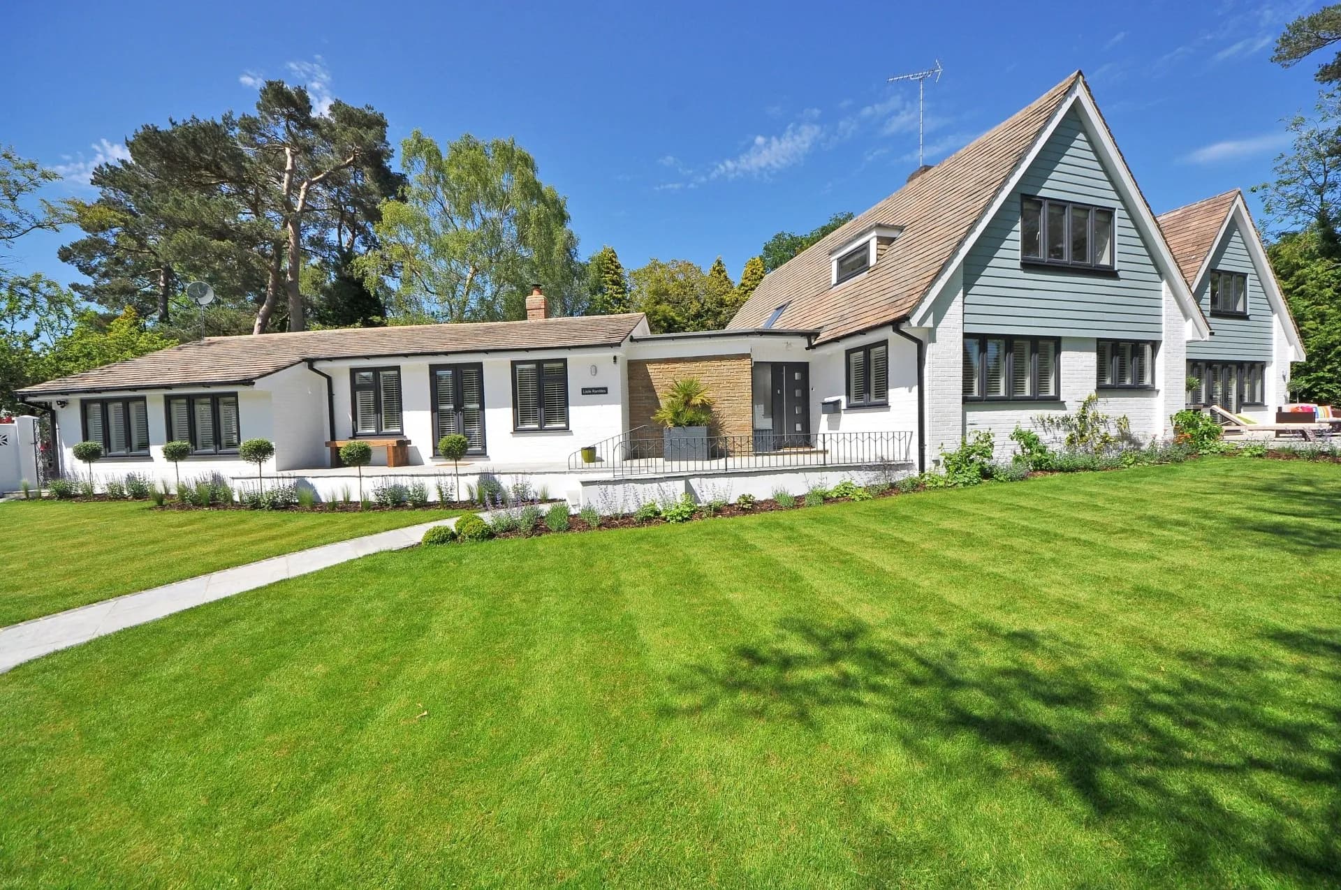 White house with grey-blue siding and two gabled roofs, windows, manicured lawn and trees under blue sky