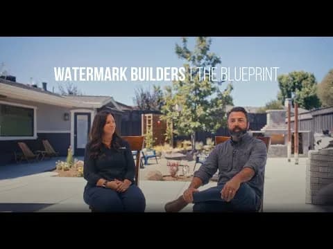 Two people sit on outdoor chairs in a sunny backyard; overlay text Watermark Builders | The Blueprint