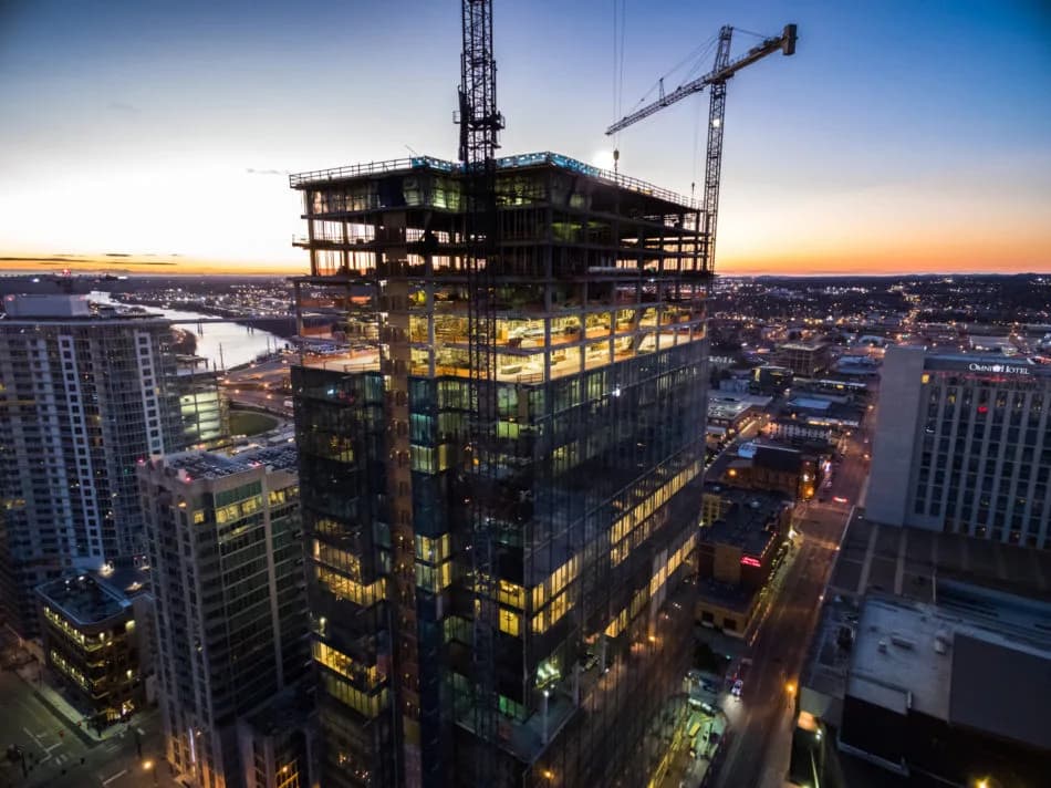 High-rise building under construction with cranes atop, sunset cityscape in the background