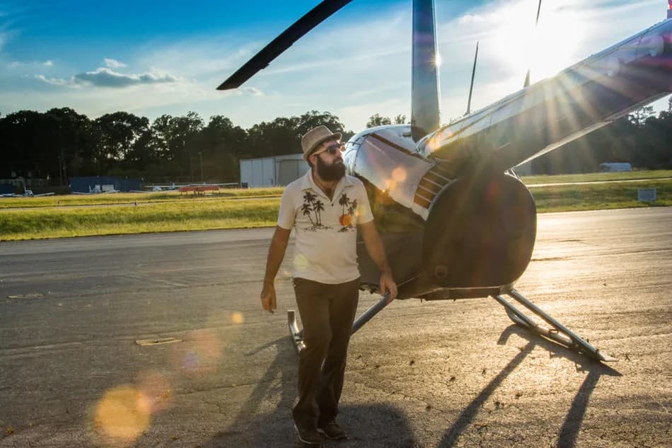 Man in a tropical shirt and hat stands beside a small helicopter drone on a sunlit tarmac with lens flare