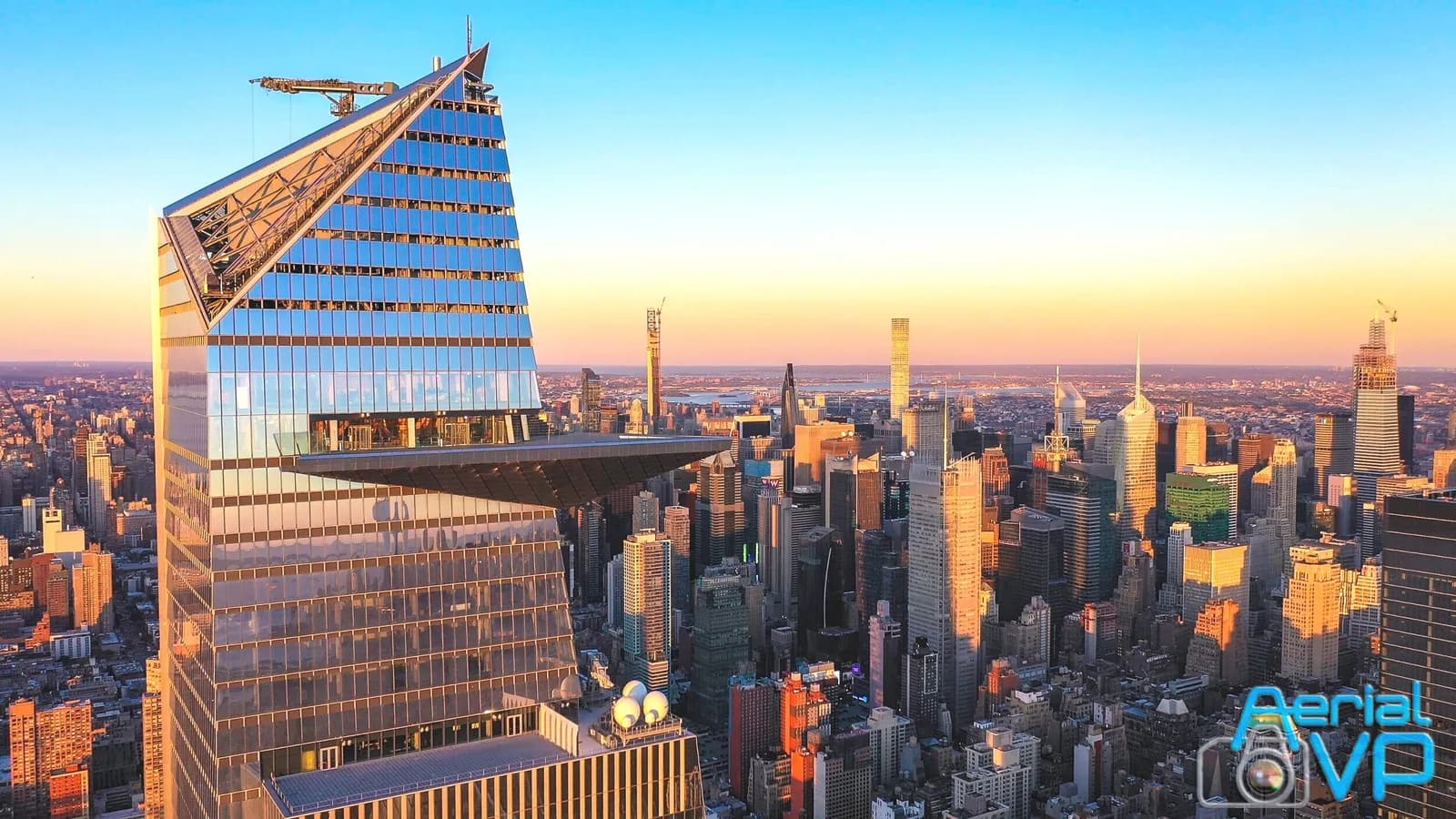 Sunset city skyline with a glass skyscraper and triangular overhang; Aerial VP logo in bottom right
