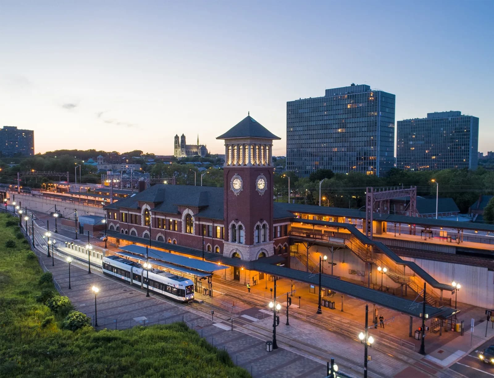 Aerial view of a brick clock tower at a train station with a white train and modern glass high-rises at dusk