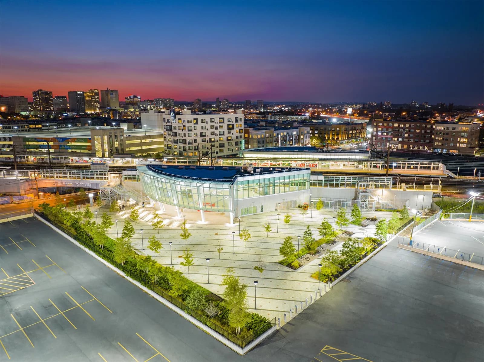 Aerial view of a curved glass building in a plaza with trees and parking, city skyline at dusk