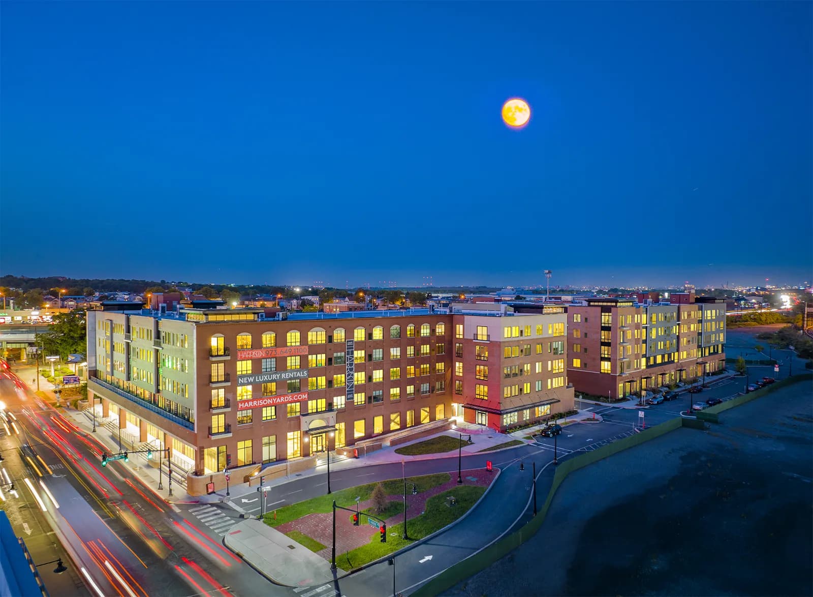 Aerial view of brick buildings with lit windows, curved road and light trails beside river under full moon