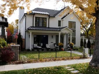Two-story white house with dark roof and trim, front porch seating, black metal fence, autumn trees