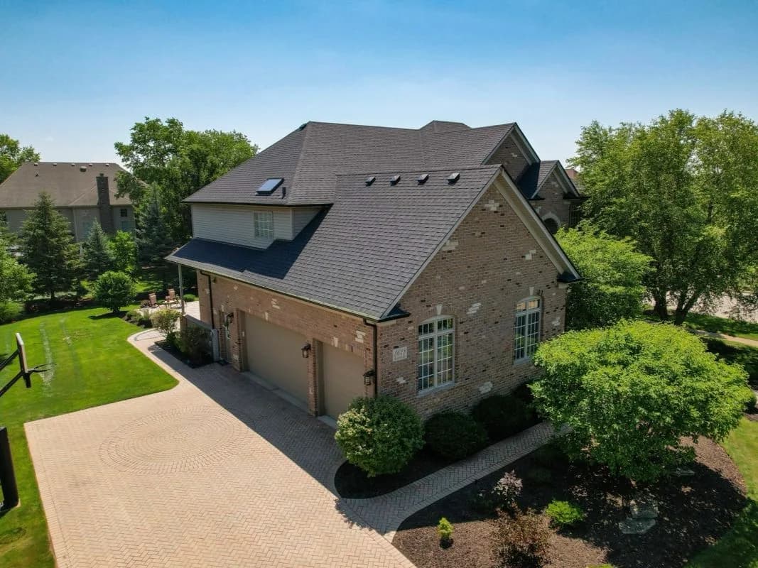 Aerial view of a brick house with a gray shingled roof, dormers, a two-car garage, and a landscaped yard