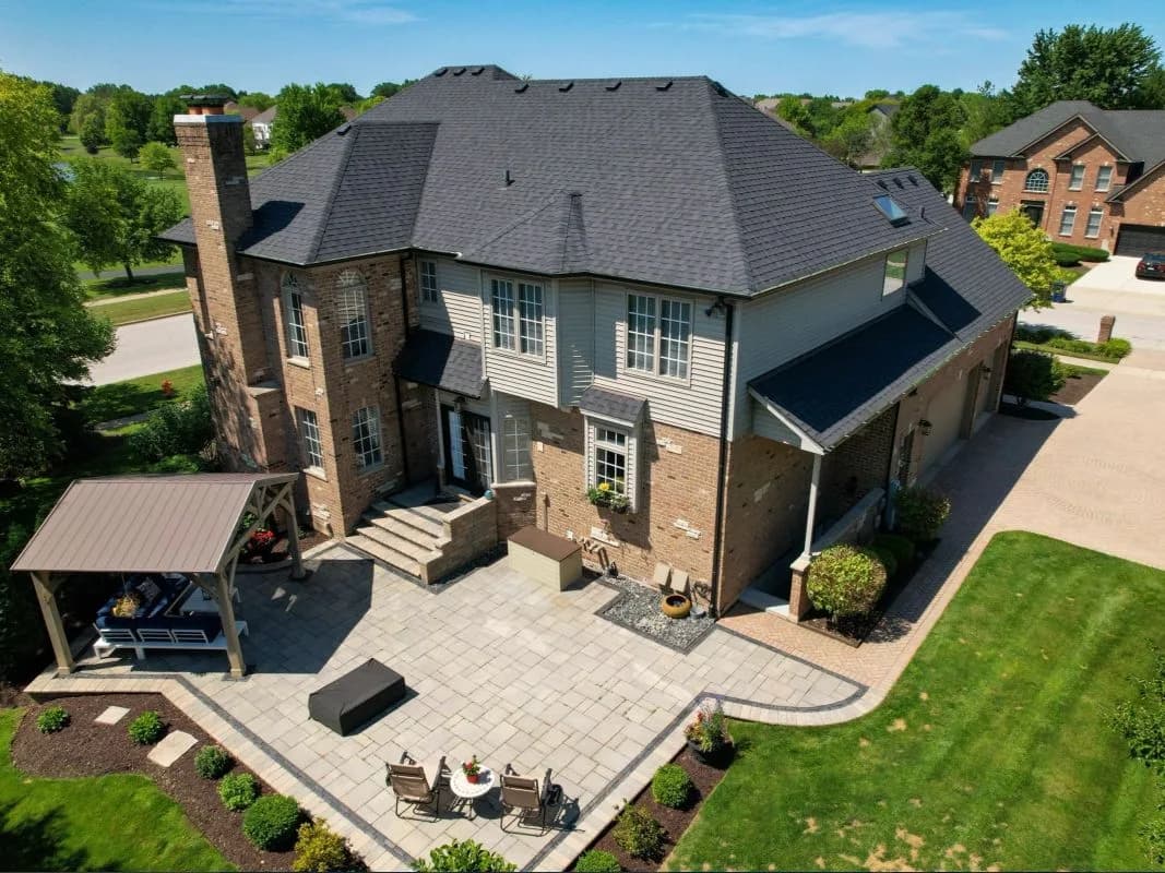 Aerial view of a two-story brick house with a gray roof, paved patio, seating area and manicured lawn