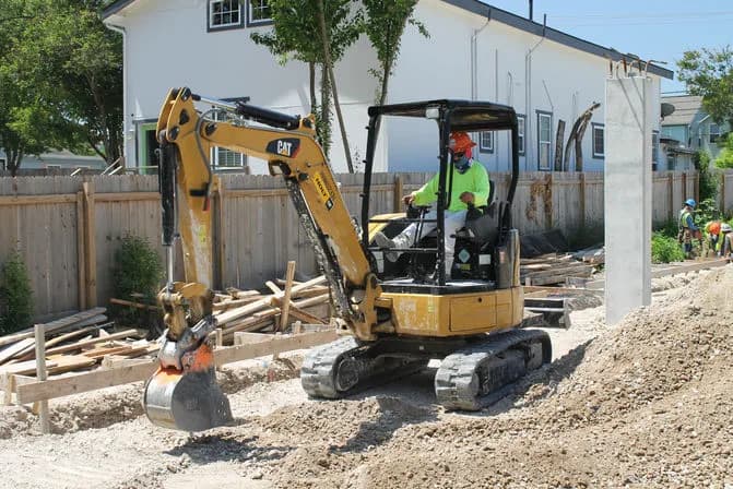 Yellow CAT mini excavator with operator in neon shirt and hard hat at a construction site