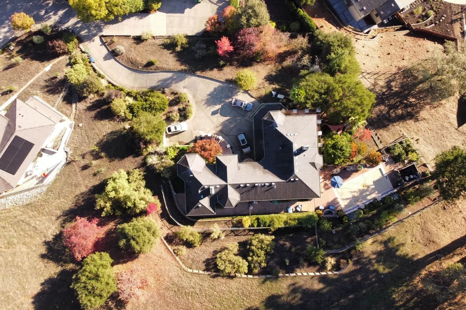 Aerial view of a large house with a dark roof, a curved driveway, cars, and surrounding trees