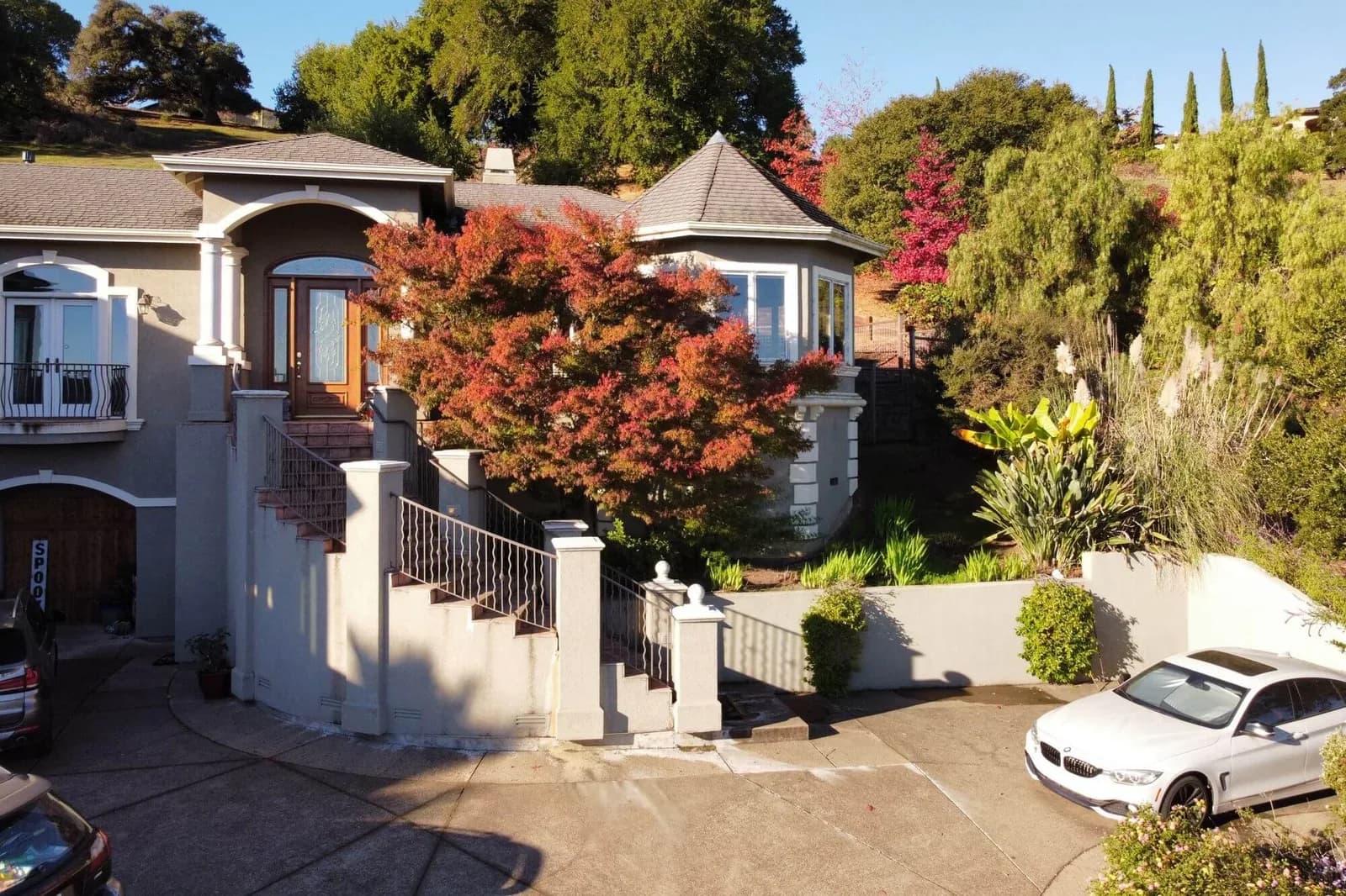 Aerial view of a beige house with arched entry, front stairs, and a red maple in yard; white car in driveway