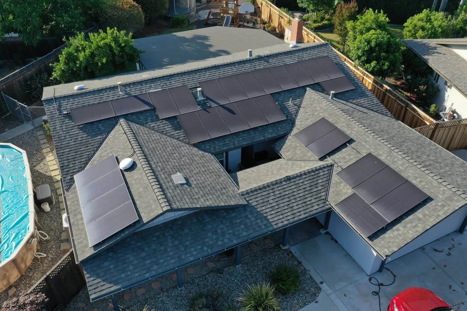 Aerial view of a multi-section house roof covered with solar panels, with a blue pool nearby