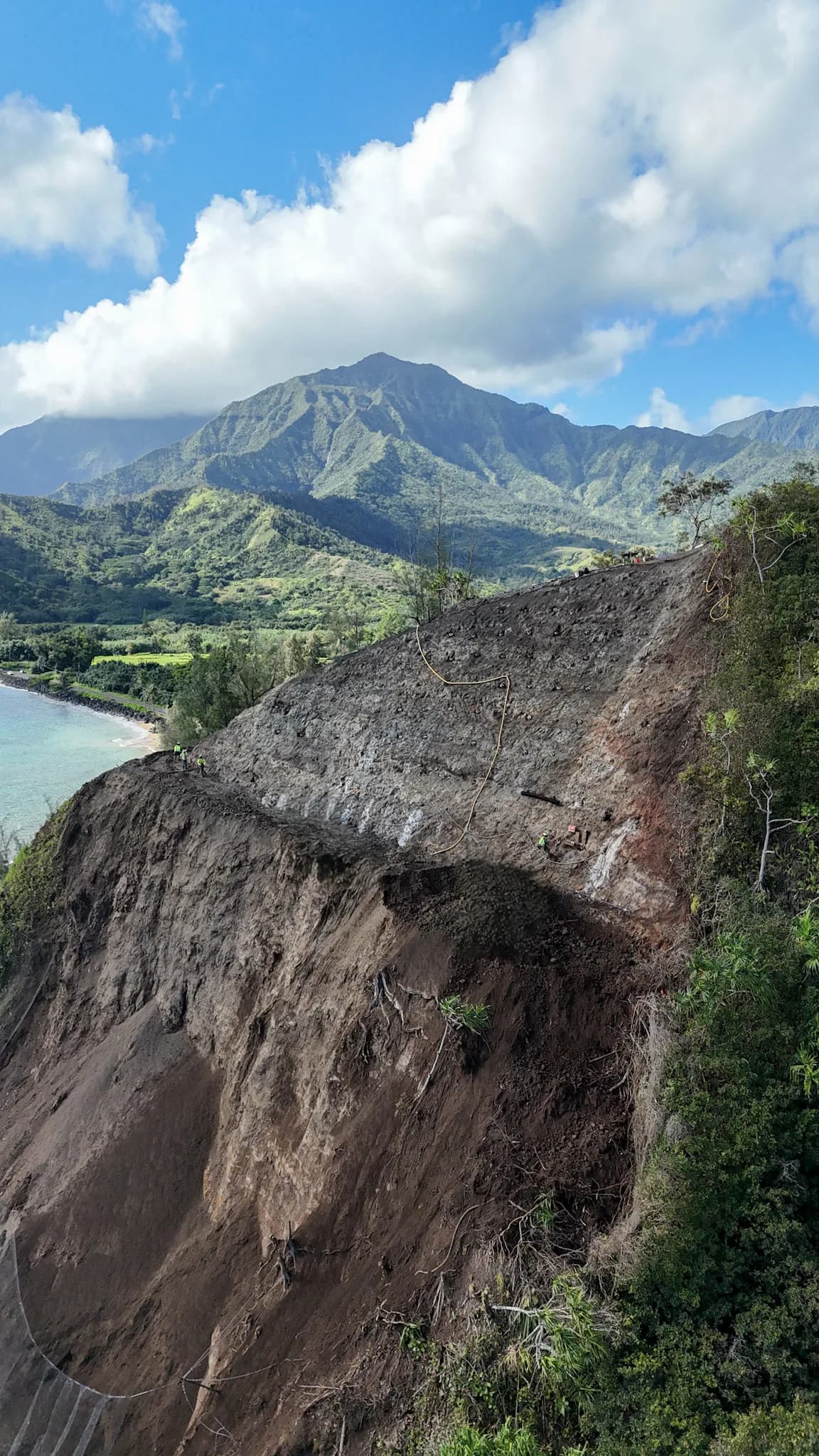 Aerial view of a steep brown cliff by the coast, turquoise water to the left and green hills beyond