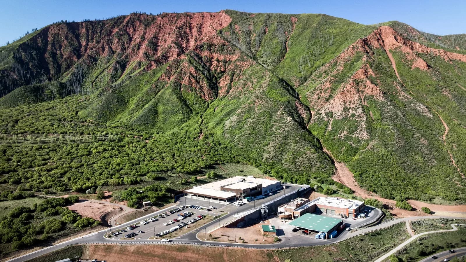 Aerial view of green hills with red rock seams and a building complex with a parking lot