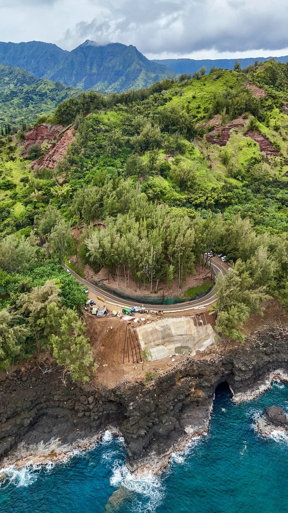 Aerial view of green hills and rocky cliff, with a curved road and turquoise water below
