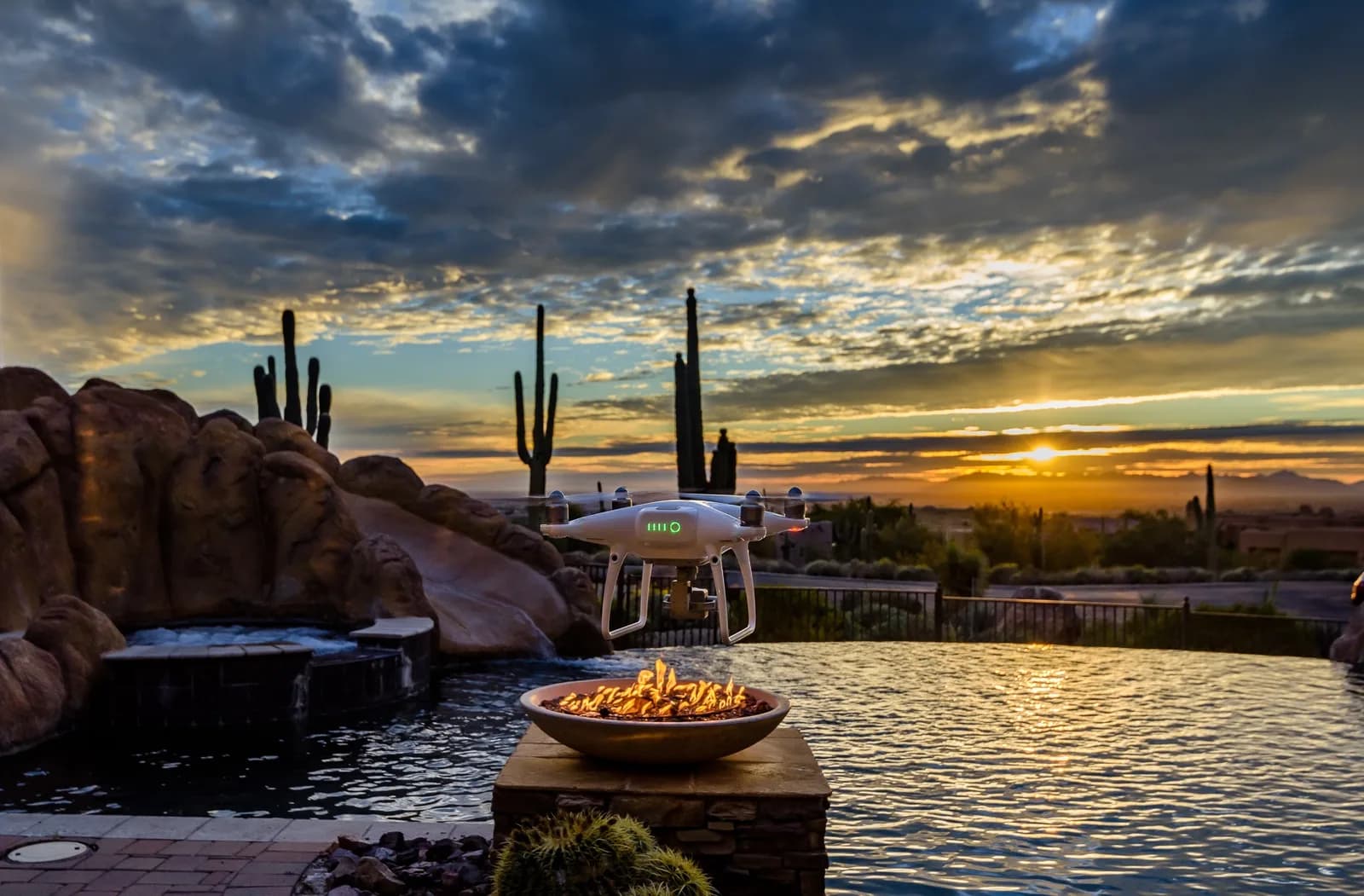 White quadcopter hovering over a lit fire bowl beside a pool at sunset with saguaro cacti