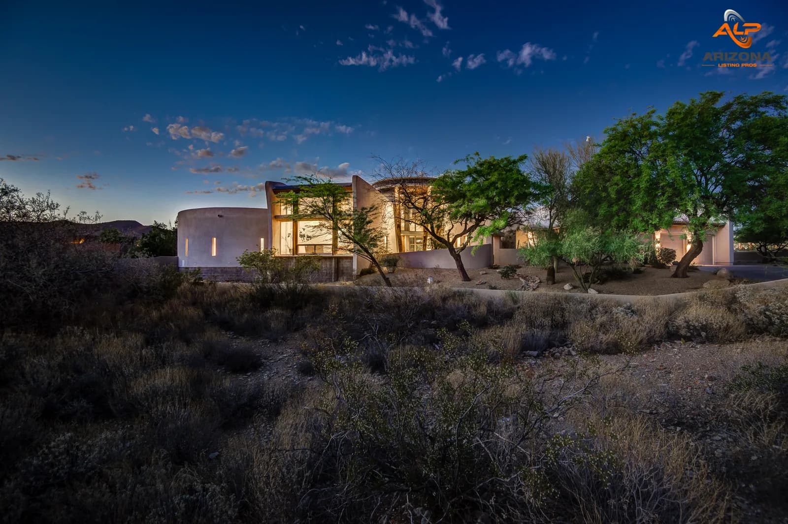 Twilight exterior of illuminated modern southwestern house amid desert scrub and trees