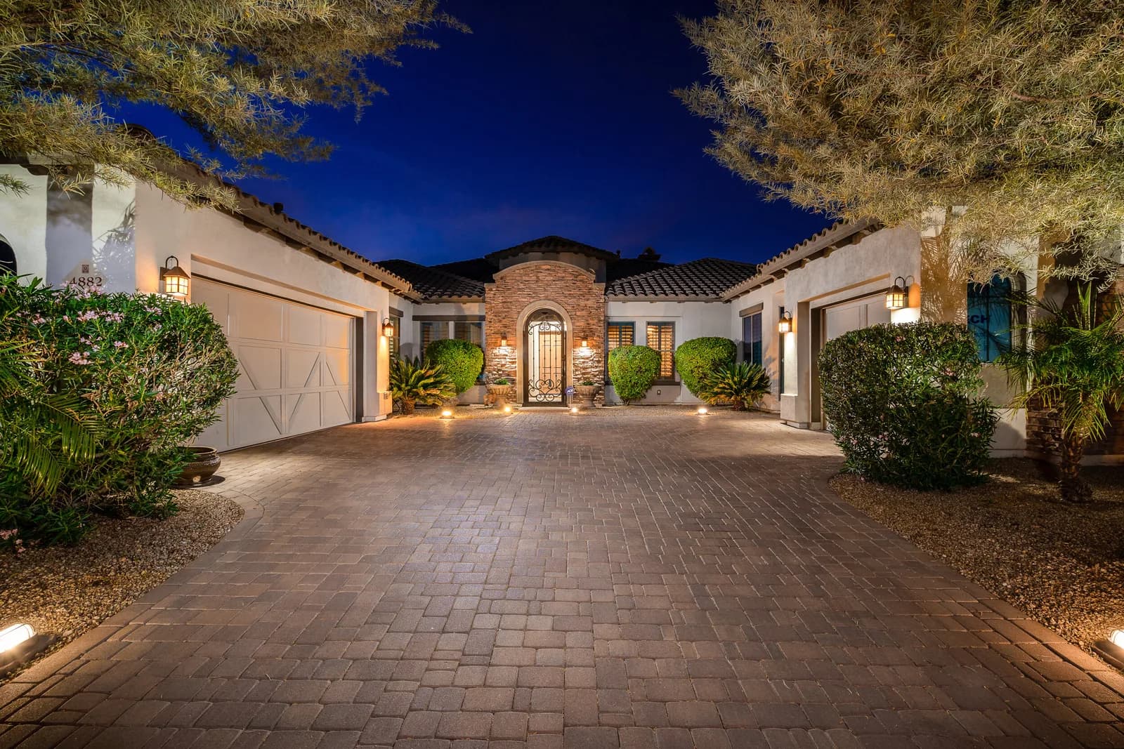 Lit Mediterranean-style house with arched iron entry and paved driveway at dusk