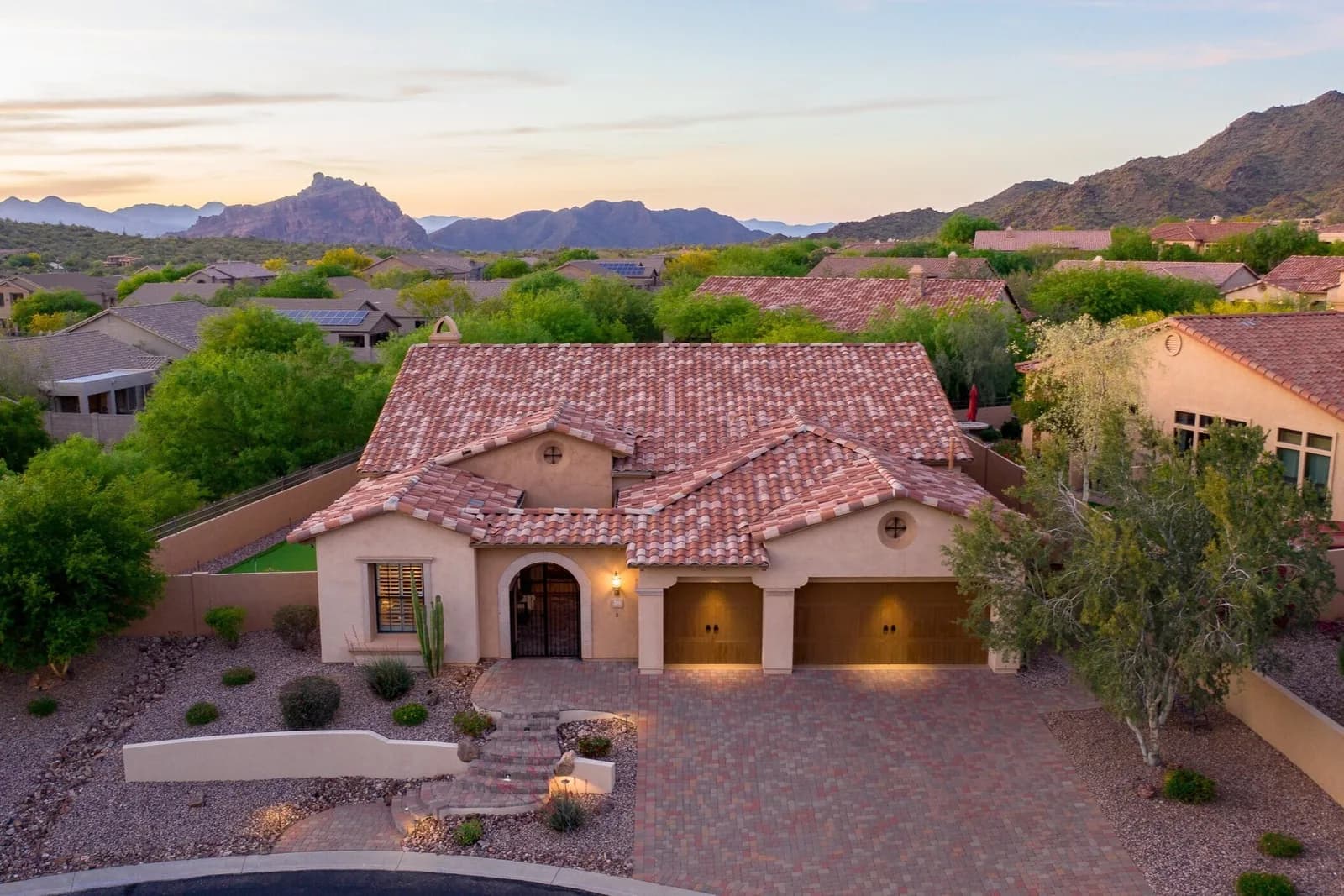 Tile-roofed suburban house with three-car garage viewed from above at sunset, surrounding homes and mountains