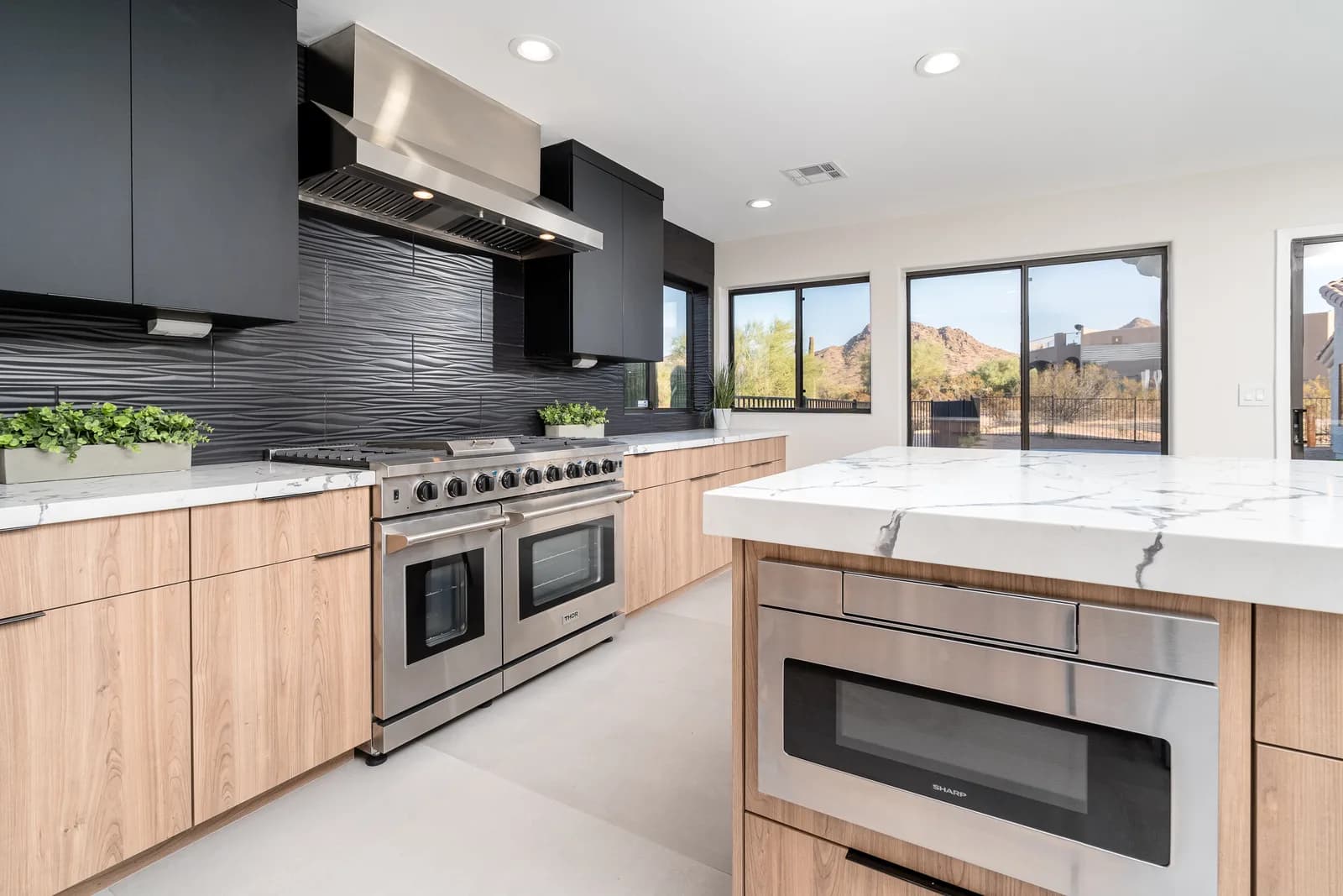 Modern kitchen with stainless steel double oven, marble island, wood cabinets and desert view through windows